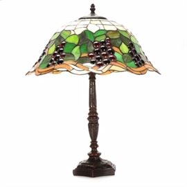 Reproduction Tiffany Style Table Lamp: A reproduction Tiffany style table lamp. This one-fixture lamp has a stained glass style, rounded bell shade and is decorated with green leaf and purple grape cluster beads. The candlestick base has fluted column detailing, an in-line cord switch, and is finished with a decorative, pointed finial.