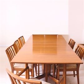 Danish Modern Dining Table and Chairs: A vintage Danish Modern style teak dining set. This dining table features sleek surfaces, collapsible drop-leaf ends, and a total of three insertable leaves which can expand the table to 105" long. The movable, spindled, wide legs allow the ends of the table to drop which can condense the table to only 25" in depth. Included with this table are six chairs, one of which is a captain’s chair with arms. Each chair has a cream colored, textured fabric cushion and spindled back with clean angular lines and square tapered legs. Unmarked.