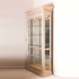 Painted Wood and Glass Display Cabinet: A glass display cabinet with wooden frame. This cabinet has a wooden frame which has been painted a cream color and has a carved decorative border around both the top and bottom of the frame. The cabinet has a glass front and mirrored interior back with four hinged side doors for ease of placing items onto any of the four adjustable glass shelves. The cabinet also includes interior overhead lighting.