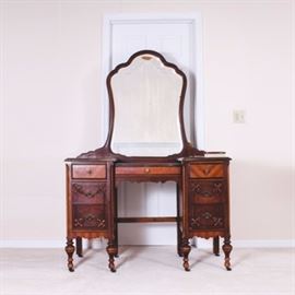 Wooden Vintage Vanity with Mirror: A vintage wooden vanity with mirror. This vanity is composed of dark stained wood and supported with a total of eight legs with wheels. Below three simple drawers are four drawers, equal in size, elaborately carved with fleur de lis. A mirror sits atop the desk surface of the vanity. Also included is a partial vanity set which includes a handheld mirror and two brushes.