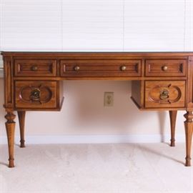 Vintage Wooden Desk: A vintage wooden desk. This desk has been stained a rich chestnut color and has a total of five drawers. A protective, movable glass top sits on the desk surface and each drawer has decorative carving with the bottom two drawers having a flower shaped mount with bail handle. Stands on four tapered legs.