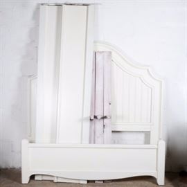 White Painted Bedstead By Howard Miller: A white painted bedstead by Howard Miller. This bedstead with an arched headboard with applied moldings around two inset panels of vertical tongue and groove boards has a painted white finish; with two wide side rails with applied moldings, three splats with center leg supports and a matching foot board with applied molding at the top and at the scrolled bottom; all resting on the four square corner posts. Marked on headboard: Howard Miller Signature Home Furnishings by Ty Pennington.