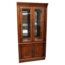Thomasville Walnut Illuminated Cabinet: A Thomasville walnut illuminated cabinet. This rectangular top cabinet comprised of English walnut with distressed finish and molded trim features two cabinet doors with clear glass panels and brass handles opening to two clear glass shelves, a mirrored back panel, and recessed lighting. The lower base features two cabinet doors with recessed panels and terminates on a plinth base. For matching pieces, see item 17IND178-172, 17IND178-142, 17IND178-154, and 17IND178-128.