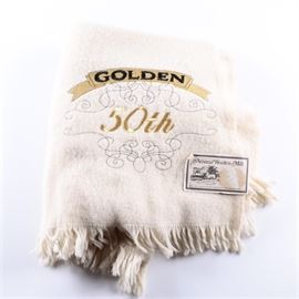 Amana Woolen Mill Golden Anniversary Blanket: An Amana Woolen Mill Golden Anniversary blanket. The white wool blanket features a rectangular form with fringe to the edges. It reads “Golden 50th” to one side in golden threads with grey scroll accents. It was made in Iowa and is marked “Amana Woolen Mill” to the tag.