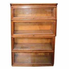 Vintage Barrister Bookcase: A vintage barrister bookcase. This four piece unit features a walnut stain and rectangular top with four glass pane cabinet doors. The cabinet doors lift up and feature a brass knob handle. This piece sits on a plinth base. There are no visible maker’s marks.