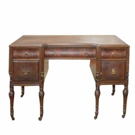Vintage Sheraton Style Walnut Writing Desk: A vintage Sheraton style writing desk in walnut, having a shaped top with molded edge, above a center drawer, flanked by four drawers in flame veneered walnut, above six turned legs with reeded stiles, terminating on casters. Circa 1940s.