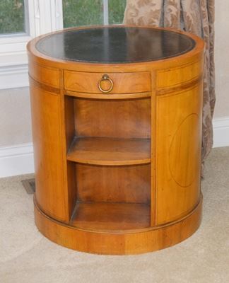 Baker Furniture Bookcase Round Drum Side Table: A round wood bookcase drum table by Baker Furniture. With a black leather inlay with embossed leaf edge, this table features three sections of two shelves separated by medallion side panels, with one center drawer. The wood is thought to be fruitwood. The drawer is marked “Baker Furniture.”