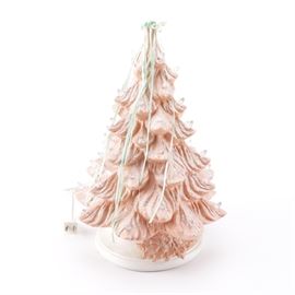 Pink Ceramic Christmas Tree: A pink ceramic Christmas tree. This piece has a glittery pink finish, with clear lights to the tips. Pale green ribbons cascade from a cream colored fabric topper. The tree stands on a white circular base with a painted pink poinsettia accent and a light socket. It is untested. The tree is marked “JS/93”.
