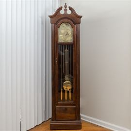 Viking Clock Company Grandfather Clock: A vintage grandfather clock by Viking Clock Company with a hand-crafted case. The clock features a wooden case that is stained a medium cherry color. The hood features a broken arch pediment with center finial and glazed glass front door below flanked by ribbed columns. The face is gold tone in color with Arabic numbers and features an hour and minute hand. An arched area above the clock face depicts the moon phases. The case has a glass front door that opens to reveal a brass lyre pendulum with three brass cased weights. The works are marked “86/Made for /Viking Clock Company/ By Hermle Black Forest Clock./451-050H/94cm/66”A winding key is included.