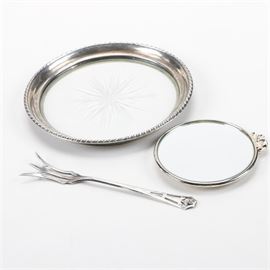Webster Co. Sterling Silver Tableware with Sterling Framed Mirror: A three-piece group of sterling silver pieces. This includes a lemon fork with pierced handle and a glass wine bottle coaster with gadroon sterling rim by Webster Co. They are both marked “Sterling” and “WCo”. The pairing is presented with a small sterling silver framed mirror that is round in shape and has a bow accent.