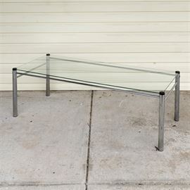 Contemporary Coffee Table: A contemporary coffee table. This table features a rectangular glass top that rises above cylindrical metal legs connected by a metal box stretcher. The table is unmarked.