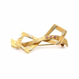 18K Yellow Gold Ruby Bow Brooch With 18K White Gold Accents: An 18K yellow gold ruby bow brooch with 18K white gold accents. This brooch features an openwork bow motif, which has a Florentine finish and three ruby accents. There is a leaf motif detail in white gold applied to the outer edge.