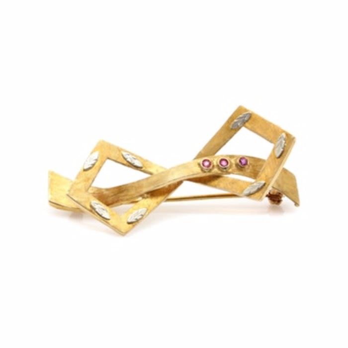 18K Yellow Gold Ruby Bow Brooch With 18K White Gold Accents: An 18K yellow gold ruby bow brooch with 18K white gold accents. This brooch features an openwork bow motif, which has a Florentine finish and three ruby accents. There is a leaf motif detail in white gold applied to the outer edge.