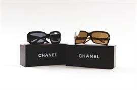 Pair of Chanel Sunglasses: A pair of Chanel sunglasses presented with their original boxes and cases. The first in the pair has a black frame and black tinted lenses with a synthetic mother-of-pearl Chanel logo inlay on the side of either arm. Interior stamps read “RAJ1920AA”, “120”, “RC016” and “Made in Italy”. The second pair of sunglasses in this pair has a brown tortoise shell frame with sepia tinted lenses and brass colored Chanel logos on either center end of the lense frames. Interior stamps read “5065”, “C502/73”, “120”, “Chanel” and “Made in Italy”.