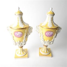 Pair of Carl Thieme Saxonian Porcelain Factory Urns: A pair of yellow Neoclassical porcelain covered urns, marked by the Carl Thieme Saxonian Porcelain Factory. Each lid features artichoke finials and acanthus leaves in bisque porcelain, on a yellow glazed ground. The main body has an oval panel with hand-painted scenes to each side, in magenta and embellished with bisque masks and floral garlands, on a yellow glazed ground with stippled cell pattern, above bolted fluted bases and a square plinth. Early 20th century.