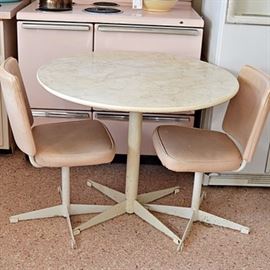 Mid Century Dining Table and Chairs: A mid century dining table and pair of chairs. The round table has a cream formica top with gold tone veins and cream edge, with a round pedestal column with four-arm base. The matching armless chairs are upholstered in a pale pink vinyl, with a cream column pedestal and four-arm base.