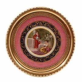 Royal Vienna Framed Hand Painted Porcelain Plate: A Royal Vienna hand painted porcelain plate depicting two classical females in a courtyard scene. One figure plays the harp while the other watches. The plate has three concentric borders with gilt floral and embossed motifs. A blue mark is present to the underside. Presented in a gilt and gesso wood circular frame.