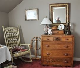 An antique wicker rocker and chest of drawers with an antique frame mirror.  I think there is a reflection of another almost antique in the mirror.