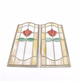 Traditional Framed Stained Glass Panels: A pair of traditional framed stained glass panels. Each panel features a brass toned metal frame with an arched crest, as well as brass toned cames. The panels also feature a single red tulip motif with a pair of green leaves set against an ivory to amber background. They both go visibly unmarked.