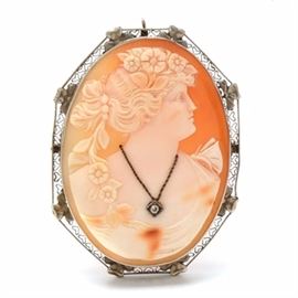 14K White Gold Shell and Diamond Cameo Brooch: A 14K white gold shell and diamond cameo brooch. This brooch features carved cameo shell, with the woman in the cameo wearing a necklace accented by a diamond, and it has a heart motif filigree border.