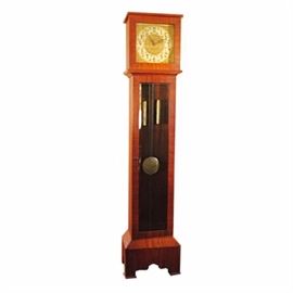 Wood Grandfather Clock: A grandfather clock. The clock has a wood case with a cherry stained finish and features a square bonnet with a hinged, glass front door. The clock has a white face with black Arabic numbers and serpentine hands on an embossed, brass floral design with raised cherub and scrollwork spandrels. The clock has a lower glass front door that displays a brass pendulum and cylinder style weights on chains and the clock stands on a tapered base with ogee bracket feet.