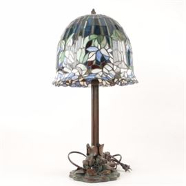 Tiffany Reproduction Bronze Tone Table Lamp With Slag Glass Shade: A Tiffany reproduction stained slag glass shade with a bronze tone metal table lamp. The bell-shaped glass shade features blue and green foliate designs. It is held by a bronze tone finial. The candle stick style lamp includes two sockets over a reeded column ending on a green patinaed leaf motif base.