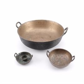 Vintage Metal Cookware: A collection of vintage metal cookware. Included are three circular pans with handles. The larger pan has a brass-tone interior and black exterior. The smaller two pans have a copper or brass appearance. There is a small metal cup with two holes in the side and one in the bottom. No visible maker’s marks are present.