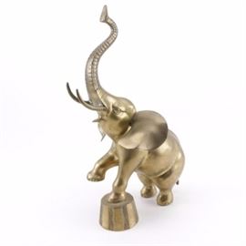 Brass-Tone Metal Elephant Sculpture: A brass-tone metal sculpture of an elephant. This sculpture depicts an elephant with an upturned trunk balancing one front leg on a circus style stand. It is labeled with a sticker that reads “made in Korea”.