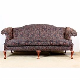 Chippendale Sofa by Woodmark: A Chippendale style camel back sofa by Woodmark. This piece comprises an arched crestrail, rolled arms, one loose seat cushion and tailored apron. It stands on front carved cabriole style legs with pad feet. It is upholstered in a jewel tone paisley fabric. A maker’s tag is seen under the cushion.