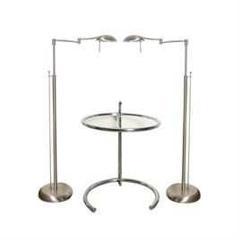 Contemporary Side Table with Two Floor Lamps: A contemporary side table with two floor lamps. This selection includes one side table with a silver tone metal frame and round glass top; two matching silver tone task floor lamps with a swing arm, and a silver tone dome shade with one light socket. The lamps have a silver tone adjustable pole body on a round base.
