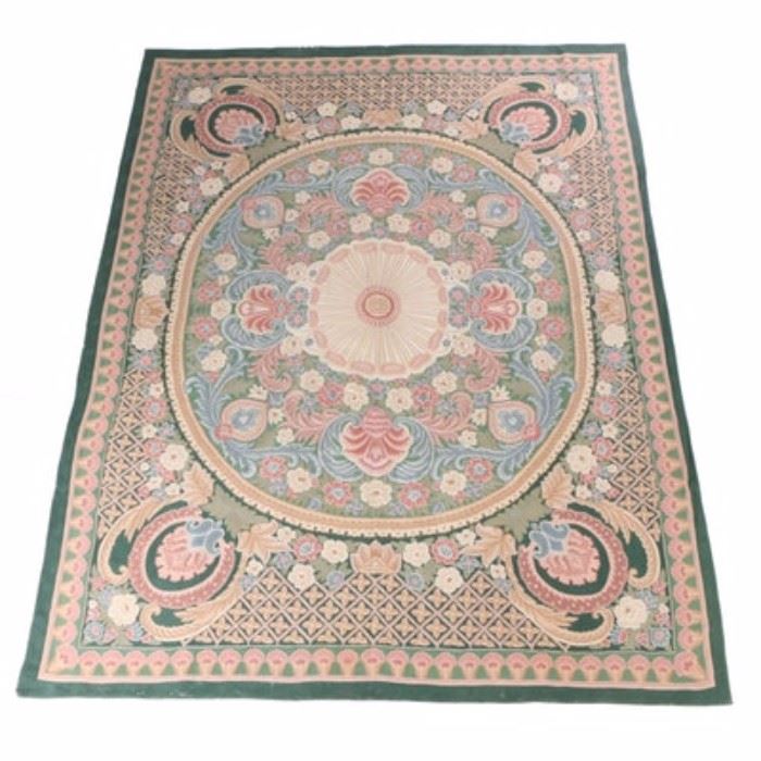 Handwoven Indian Wool Area Rug: A handwoven Indian area rug. This wool rug is chain-stitched in a palette of rose, sage, slate blue, beige, celadon, cream, forest green, and dusty pink. It features a neoclassical design beginning with an oval inner field with a floret medallion at the center amid palmettes, scrolling acanthus, flowers, and leaves. The inner field rests on a rectangular outer field, which is grounded in forest green and populated with a combination of diamond lattice patterns, scrolling acanthus, and palmettes cameos. The field is framed by compound borders including a primary border grounded in celadon and filled with pink and cream palmettes. Edges are serged on all sides under a full fabric backing. The rug is unlabeled.