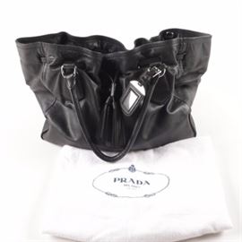 Prada Leather Drawstring Tassel Tote Bag: A Prada leather drawstring tassel tote bag. This black bag features a drawstring closure with oversized tassel detailing to the front, silver tone pierced logo, and leather handles. The interior is lined in a logo jacquard and marked with silver tone black enameled tag. Includes original dust bag and authenticity cards.