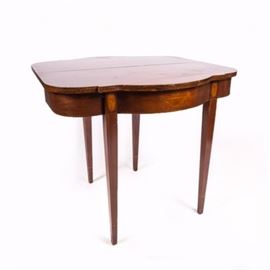 Heppelwhite Style Mahogany Game Table with Marquetry from Fine Arts Furniture: A Hepplewhite style mahogany game table with marquetry having a shaped top and inlaid edge over a compliant apron with a swelled front with an urn marquetry and an inlaid edge. The table stands on four square tapering legs, two of which are topped with oval inlays. The two back legs swing into place to support the top when it is folded open. The table has retained the original “Fine Arts Funiture Co. Grand Rapids” sticker which was in production from 1925 to 1977.