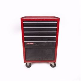 Craftsman Rolling Tool Chest: A Craftsman rolling tool chest. This piece features a panel case with recessed top over a collection of drawers including dividers, metal rails and integrated handle fronts. The piece includes a cabinet door for larger tools at the bottom, it is finished in a red tone and it includes casters with locks. The piece is marked “Craftsman”.