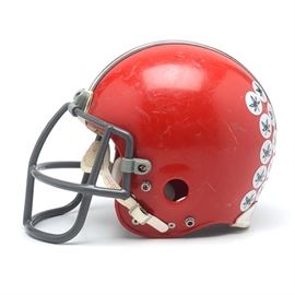 Ohio State Buckeyes Game "Throwback" Full Size Football Helmet: An Ohio State University Buckeyes 1960s full-size game “Throwback-Style” football helmet. This game issued helmet was manufactured by Riddell honors the 1968 National Championship and retains its original facemask and chin strap. There is a “Buckeyes” name plaque attached to the forehead area, just above the facemask. This helmet is painted in a scarlet color with black, white, and gray stripes that run to the front and back. With backside American Flag and NCAA decals, this helmet was obtained through the Ohio State Athletic Department according to our consignor. This helmet is adorned with Buckeye game achievement decals and maintains its original inside protective padding. It is a size 7-7.5 according to the label.