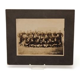 1924 "Center Mohawk" Football Photo: A 1924 football photo. The photo has “Champions 1924 Center Mohawk Merchants Foot Ball Team”; the photographer is listed, “Wood Photo 106 – 7th St.” Different merchant names are sewn on the front of the players’ jerseys; names like: Hickman Cafe, Kobmann Hardware, Kramer Meats, and Friedels Candy. One of the non players in the photo is holding a megaphone that is adorned with a “C”. Cincinnati has a Mohawk area. The photo is attached to cardboard; the names of the players are handwritten on the back.