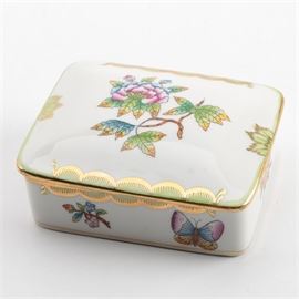 Ornate Herend Trinket Box: A Herend Hungary Queen Victoria trinket box. This ornate trinket box is made of a porcelain material and has a white finish which provides the surface for green, gold tone, pink, blue, and brown floral depictions and nature motif accents. This lidded box is marked “Herend Hungary” on the underside of the base with a maker’s mark.