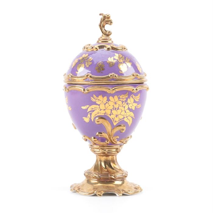 House of Faberge "Violet" Musical Egg: A vintage House of Faberge Violet musical egg. The handpainted porcelain piece was produced circa the 1980s and includes a decorative brass tone metal finial, trim and footed base. The musical egg Violet plays Tchiakovsky’s “Swan Lake”. The piece is stamped to the underside with the above information as well as “©TFM”. A label reads “Crafted in Thailand”.