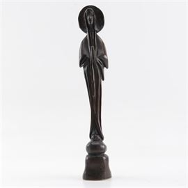 Carved Wooden Saint Figurine: A carved wooden saint figurine. This stylized figurine depicts a slender, robed figure with a halo on a pedestal in a nearly black stain. This piece is illegibly signed in black permanent marker to its underside.