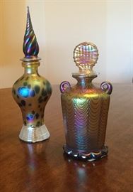 Perfume bottle in a gold finish with iridescent bubble pattern & spiral tip stopper, clear ribbed base (on left), gold and titanium perfume bottle in a cylinder shape with 2 looped handles at top, & decorative disc shape stopper, wavy iridescent colors with scallop shape base (on right)