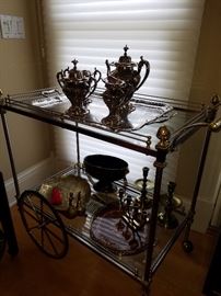 Silvered and golden accent metal tea or cocktail cart with glass upper and lower shelves displaying silverplate tea set and other metal items