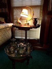 One of a pair of pedestal tables, displaying a lamp, and other items.  Foreground: Victorian tea set (silverplate) on a foot stool