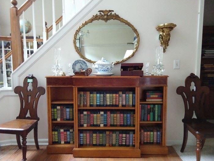 Vintage English mahogany bookcase, (measures 5' 1" L x 12 1/2" D x 40" T) with floral inlay, vintage gilt wood oval mirror, collection of leather bound books, flanked by a pair of antique English mahogany slipper chairs.                                                                                        
The wall sconce is an antique French gilt carved wood wonder!