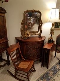 Baronial demilune cabinet with gilt frame mirror.  Press-back rocker in front. Narrow dropleaf table is rosewood