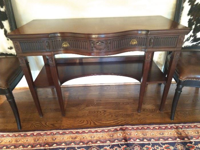 Federal style mahogany console, having a modified serpentine top, above a frieze drawer decorated with alternating fluting and parterre centering a reserve of bas relief floral swags, the whole rising on four front square tapering legs with fluting, joined by a lower shelf, measuring 31"h x 48"w x 21.5"d.