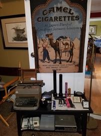 Camel Cigarettes tapestry, vintage typewriter, Mag-Light flashlights & others, electronics, small black bench/shelf.