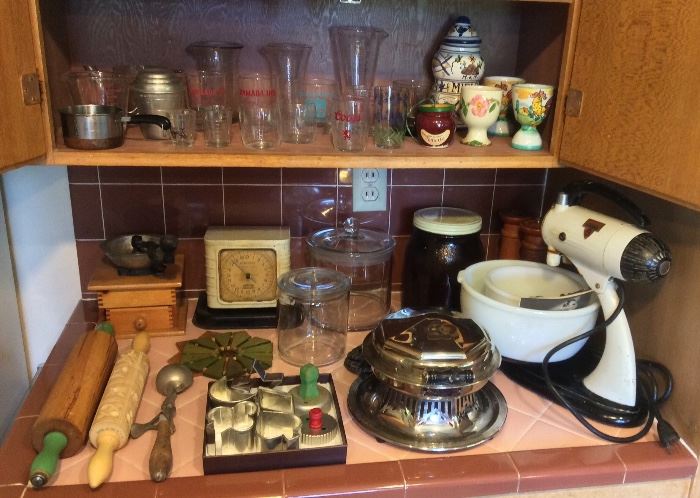 Vintage kitchen items: rolling pins, ice cream scoop, hand crank coffee grinder, cookie cutters, utility scale, glass canisters, Art Deco waffle iron, Sunbeam Mixmaster stand mixer. Above: hotel glasses, Swanky Swig, glass measuring beakers, egg cups & more