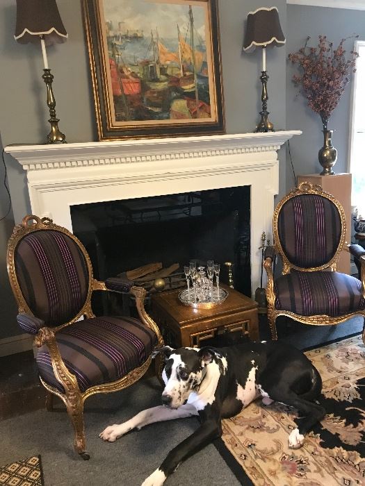 A pair of  gilded French chairs covered in  Brunswick silk, an original oil painting above the fireplace flanked by a pair of vintage lamps with newer silk shades. The Asian Table holds a  small bohemian to counter and crystal cordial glasses. Antique brass fireplace tools, fender and  andirons.