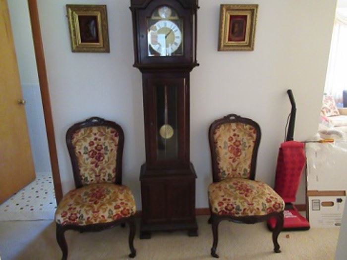 Randolf tall case clock,   flanked by walnut upholstered chairs. Above the chairs are two paintings.