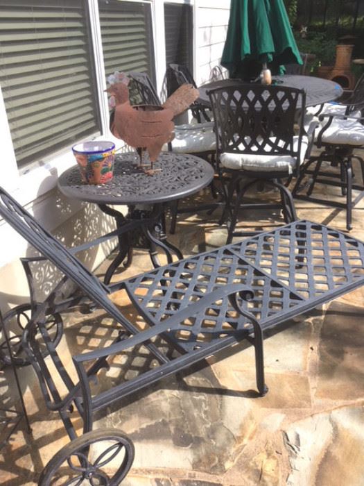 Black metal patio set includes a pair of chaise lounges, pair of small round tables, and a rectangular table with six swivel armchairs. Also a green market umbrella with metal stand. Whimsical metal rooster potted plant holder